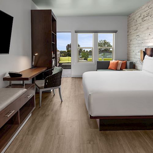 A modern hotel room with a king-sized bed, desk, TV, and a view of greenery through large windows. The decor is contemporary with wood accents.