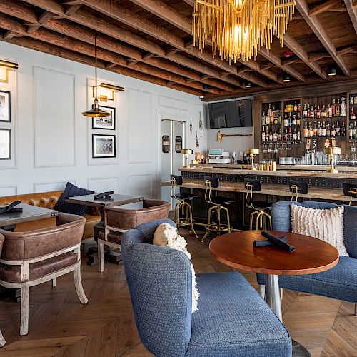 The image shows a cozy bar with wooden ceiling, plush seating, a stocked bar, framed pictures on the wall, and a warm chandelier lighting.