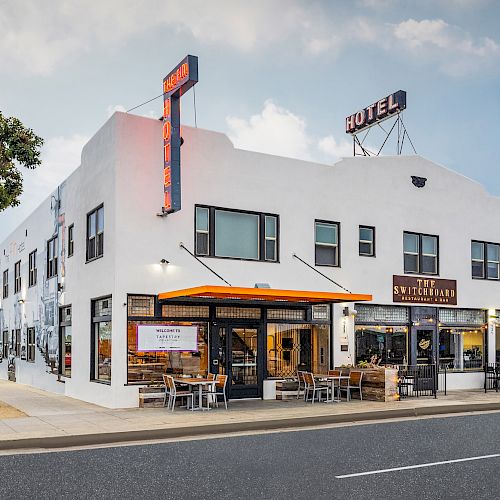 A white two-story building with a hotel sign and several shops on the ground floor, including cafes with outdoor seating, on a clear day.