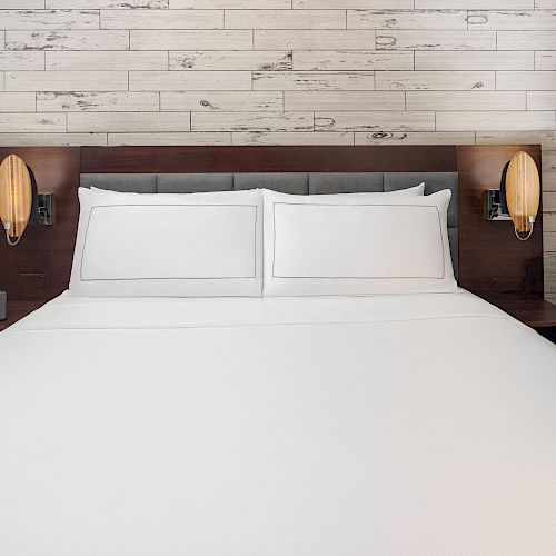 A neatly made bed with white linens against a wooden headboard and a wood-paneled wall, flanked by two wall-mounted lamps.