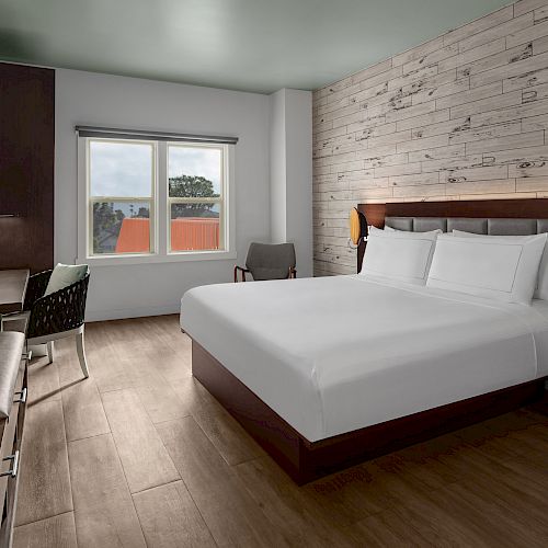 A modern hotel room features a large bed, a TV, a desk with a chair, a window with a view, and minimalistic decor with wood accents.