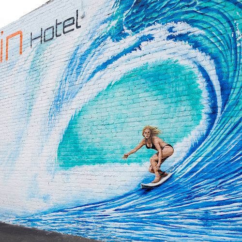 A person poses as if surfing in front of a mural of a giant wave on a wall with 