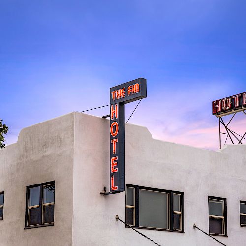 This image shows a white building with neon signs indicating it's a hotel, set against a blue and pink sky, with a tree on the left side.