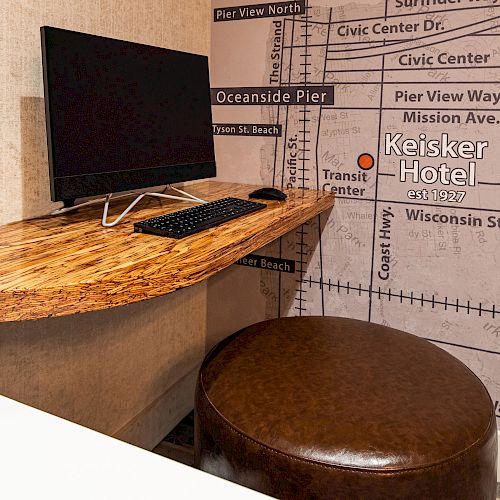 A computer on a wooden desk is positioned against a wall with a map design. An ottoman is placed under the desk in front of the computer setup.