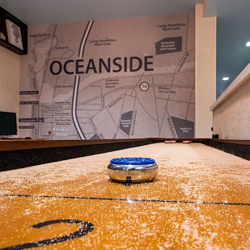 A shuffleboard table with a puck is in the foreground. The background features a map with the word 