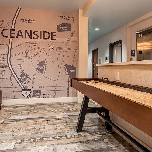 The image shows a room with a large map of Oceanside on the wall, a shuffleboard table, desk, computer, seating area, and an elevator in the background.