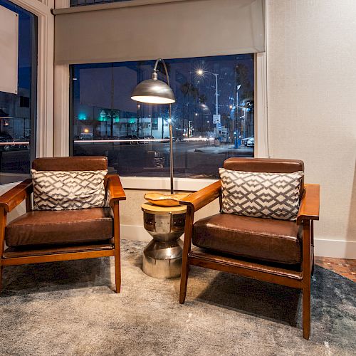 A cozy seating area with two wooden chairs, patterned cushions, a side table, and a floor lamp beside a large window at night.