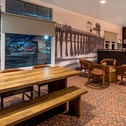 The image shows a modern, cozy café with wooden tables and chairs, featuring a large pier mural on the wall, and orange-patterned tiled flooring.