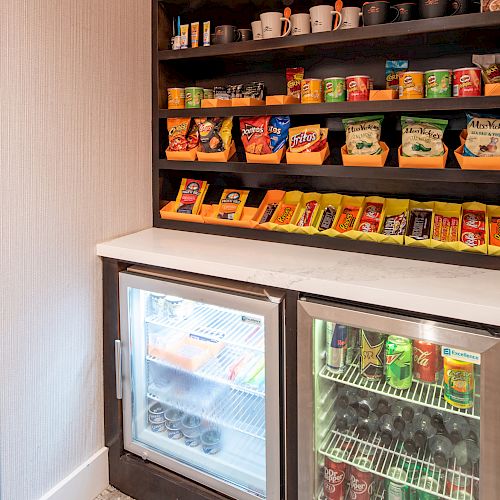 The image shows a mini snack and beverage area with shelves stocked with various chips, cup noodles, and snacks above two mini refrigerators containing drinks.