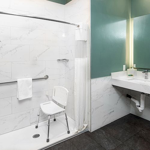 This image shows an accessible bathroom with a chair in the shower, grab bars, a sink, a large mirror, and green and white color scheme.