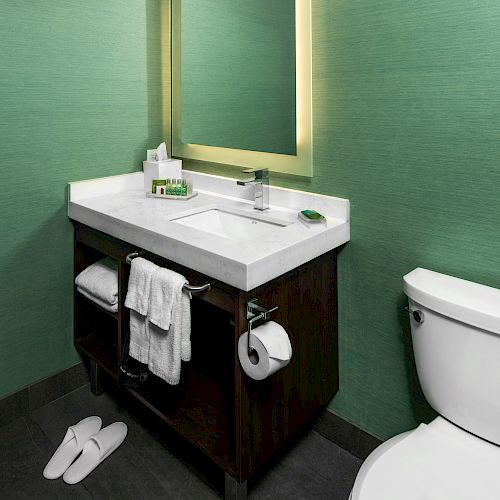 A modern bathroom with a sink, mirror, and toilet. Towels, slippers, and toiletries are visible; the walls are green.