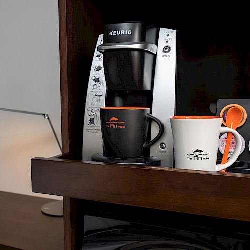 A Keurig coffee maker with two mugs and a variety of K-cups stands on a wooden surface, next to a modern desk lamp.