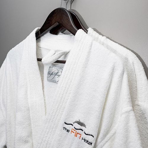 Two white bathrobes are hanging on wooden hangers against a gray wall. One has a hotel logo embroidered on it.