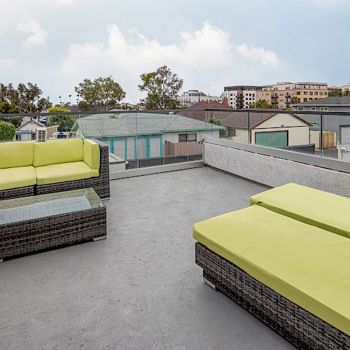 The image shows a rooftop terrace with green-cushioned lounge chairs, a table, and couches. The view includes neighboring buildings and trees.