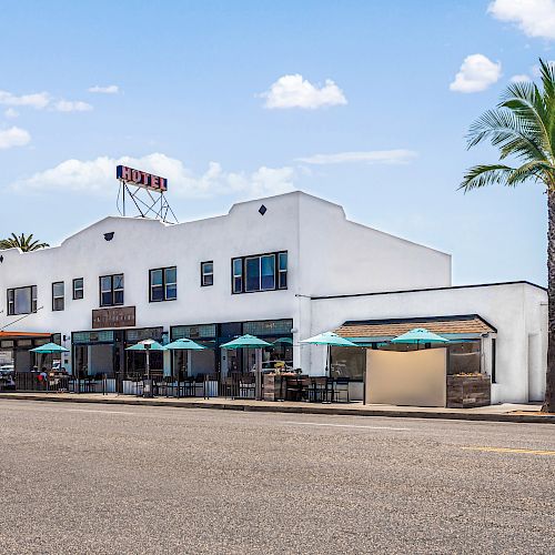 A white building with outdoor seating under umbrellas, palm trees, and a clear blue sky in the background.