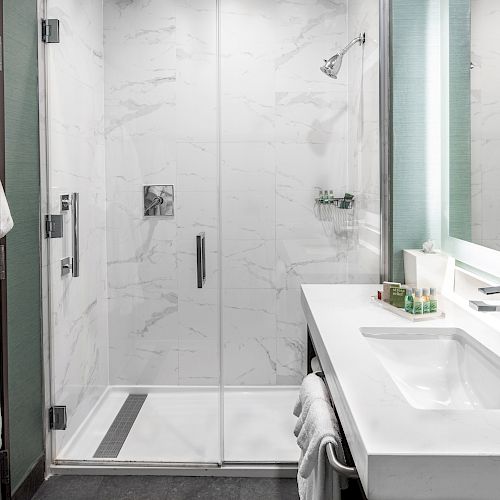 This image features a modern bathroom with a glass-enclosed shower, a white sink counter, toiletries, and a bathrobe hanging on the side.