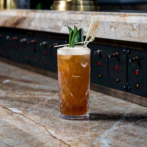 A cocktail in a tall glass garnished with pineapple leaves sits on a marbled bar counter, with a backdrop of dark panels and switches.