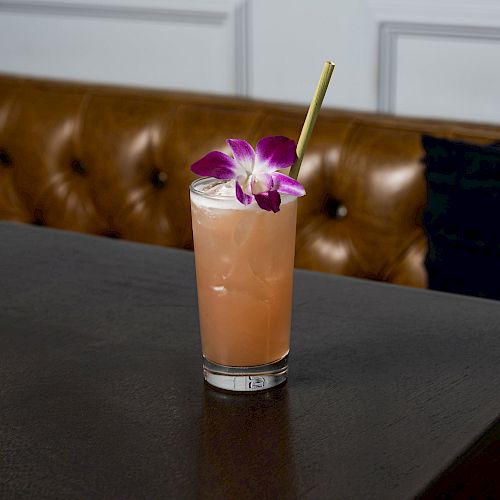 A pinkish beverage in a glass with ice, garnished with a purple flower and a straw, placed on a dark table with a brown leather bench in the background.