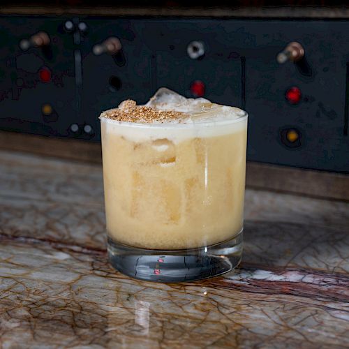 A frothy cocktail in a glass sits on a marble surface, with a control panel featuring various knobs and buttons in the background.