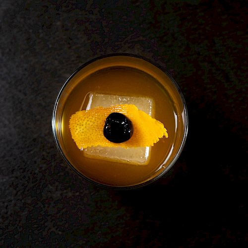 A cocktail featuring a large ice cube, garnished with an orange peel and a cherry, viewed from above on a dark background.