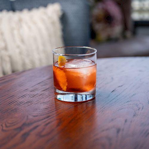 A glass with an orange drink and ice cubes sits on a polished wooden table in a cozy room with cushions in the background.