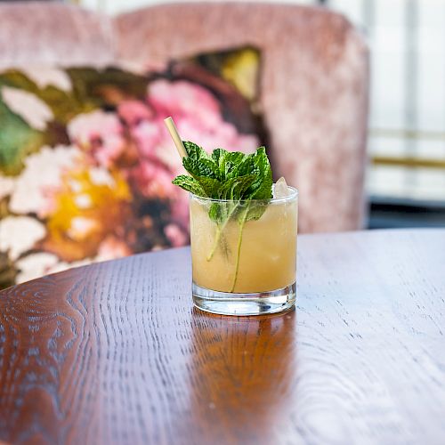 A cocktail garnished with mint leaves on a wooden table, with a floral cushion in the background.