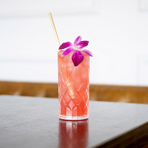 A pink beverage in a textured glass with a straw and a purple flower garnish, placed on a wooden table against a plain background.