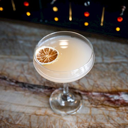 A cocktail in a coupe glass, garnished with a dried citrus slice, sits on a marbled surface with a dimly lit backdrop.