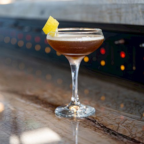 A cocktail in a coupe glass garnished with a lemon twist sits on a marble bar with blurred colorful lights in the background.