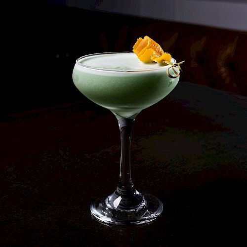 A green cocktail in a coupe glass, topped with a frothy white layer and garnished with a yellow flower, is on a dark surface.