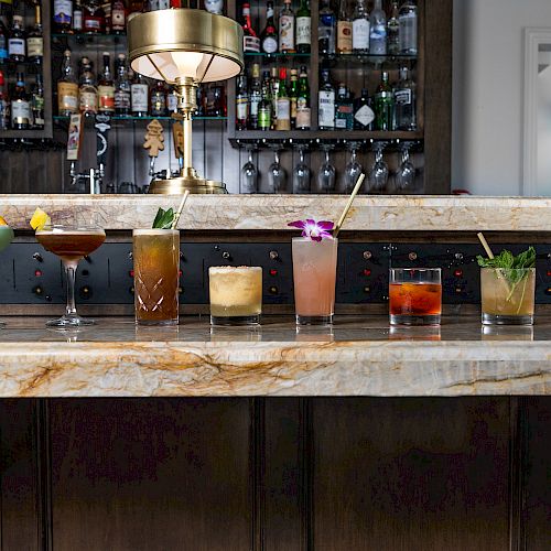 A variety of colorful cocktails are displayed on a marble bar counter, with a well-stocked bar and a golden lamp in the background.