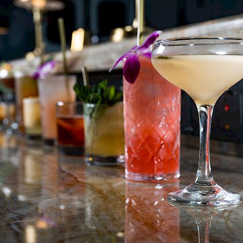 An array of colorful cocktails is lined up on a bar counter, with varying glasses and garnishes in a dimly lit setting.