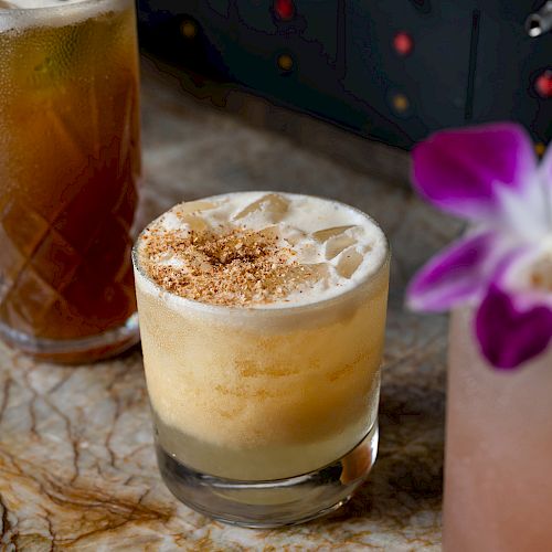 The image shows three cocktails on a marble surface, with one garnished with a purple flower and another topped with foam and sprinkled spices.