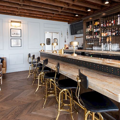 A stylish bar with modern decor, featuring a wooden counter, high chairs, shelves with bottles, and cozy seating areas in a well-lit space.