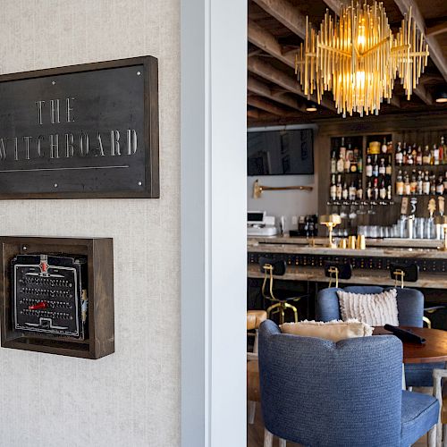 This image shows a stylish bar area with a sign labeled 'The Switchboard,' a chandelier, and seating. Bottles are neatly arranged behind the counter.