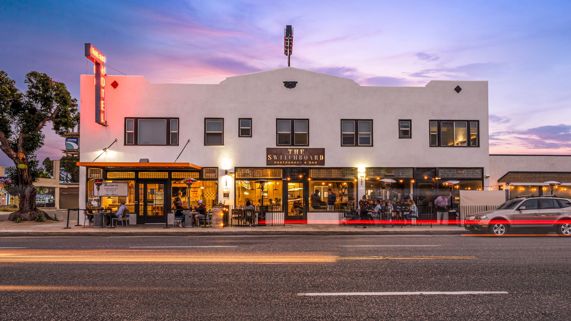 A two-story white building houses "The Switchboard" restaurant; people dine outside, and cars pass by on the street at dusk.