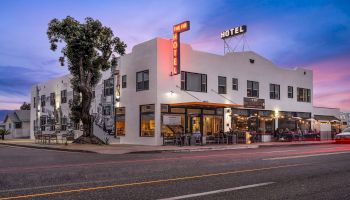 The image shows a two-story hotel building with an outdoor dining area and illuminated signs, set against a twilight sky.