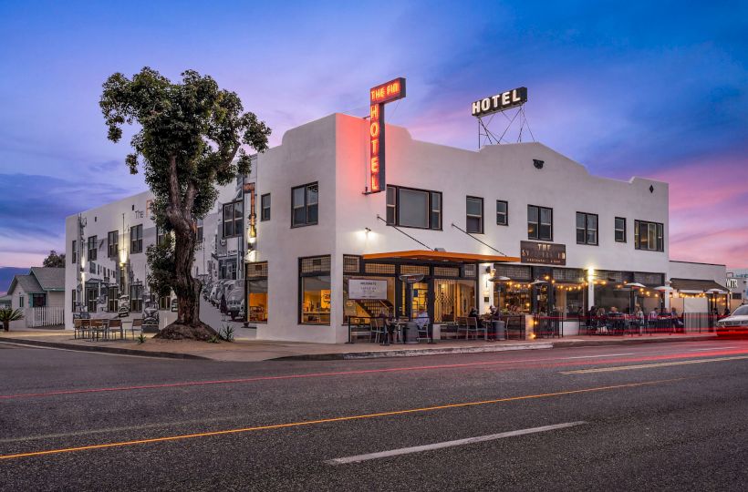 The image shows a two-story hotel building with an outdoor dining area and illuminated signs, set against a twilight sky.