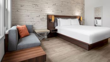 This image features a modern hotel room with a white bed, a gray sofa with a brown cushion, a large mirror, and wood-accented furniture and flooring.