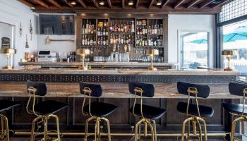 This image shows a stylish bar with a wooden design, seven bar stools, a stocked shelf of bottles, three lamps, and a mounted television.