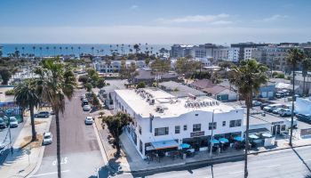 Aerial view of a coastal town with buildings, palm trees, and streets leading to the ocean on a clear day. End of description.