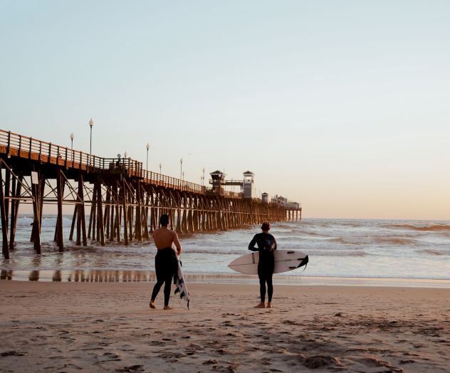 Two people with surfboards stand on a beach near a pier, with the ocean and a sunset in the background.