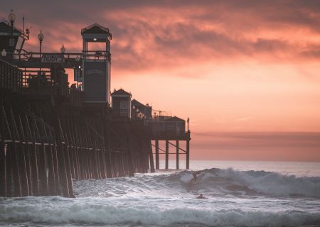 A wooden pier extends over the ocean waves at sunset, with surfboarders riding the waves beneath a dramatic, colorful sky.