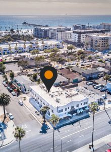 A coastal city area with a mix of buildings, palm trees, and parked cars; a location pin marks one building, with the ocean in the background.