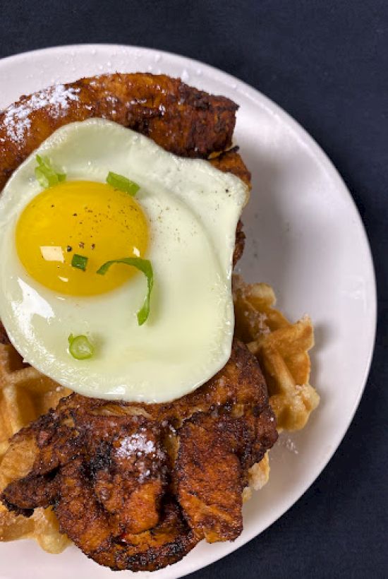 A plate of chicken and waffles topped with a sunny-side-up egg, garnished with green onions and a sprinkle of powdered sugar.