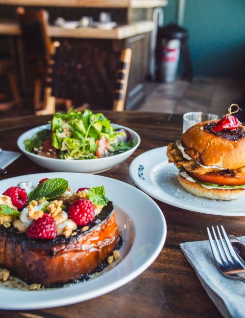 A cozy caf&eacute; table with a fresh salad, a stack of pancakes topped with syrup and berries, and a plate featuring a glossy waffle.