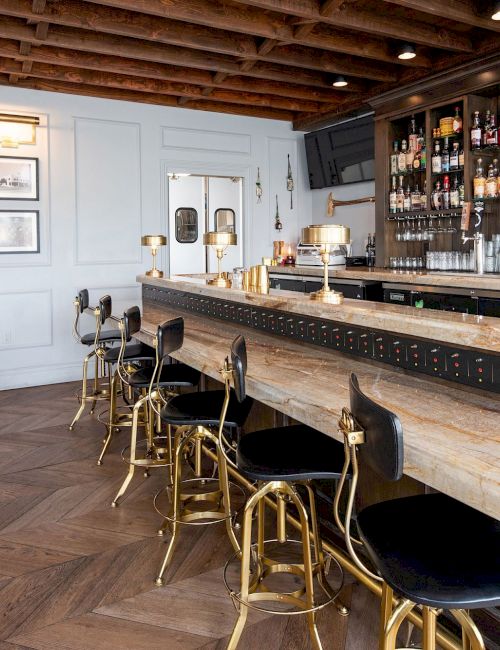 The image shows a stylish bar with wooden flooring, a countertop with bar stools, shelves of alcohol bottles, chic decor, and a seating area.