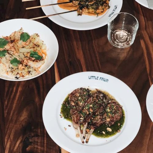 A top-down view of three plates with skewered meat and greens, plus a small cracker and a glass on a wooden table, a casual dining scene.