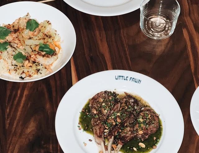 A top-down view of three plates with skewered meat and greens, plus a small cracker and a glass on a wooden table, a casual dining scene.